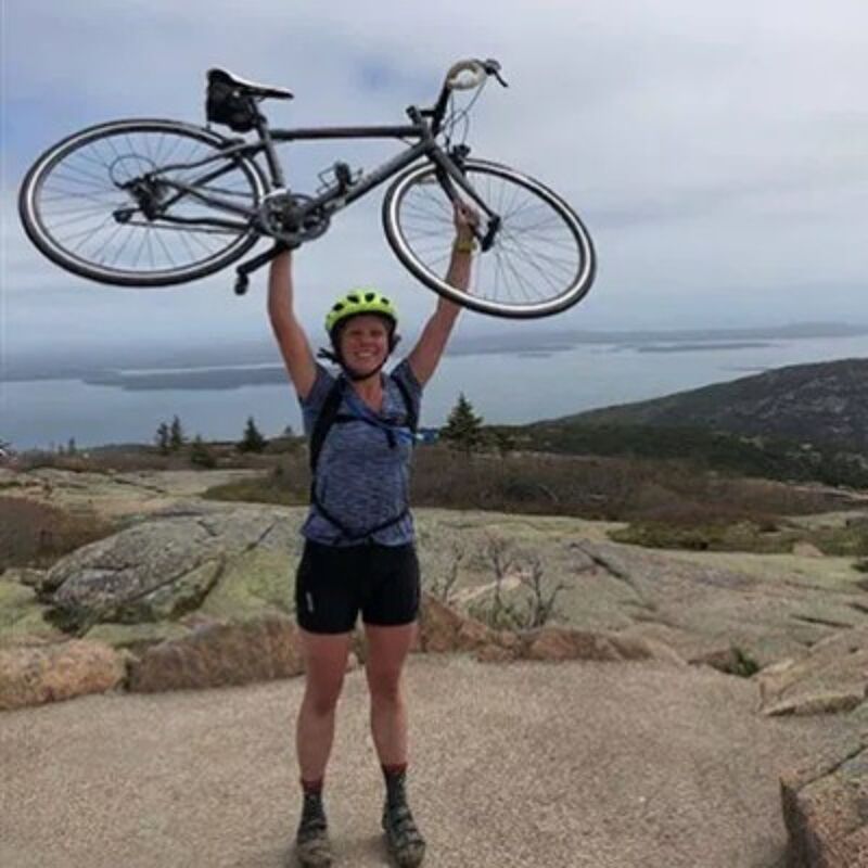 In the image, a woman stands on a rocky mountaintop, holding a bicycle above her head. She wears a helmet, shorts, and a backpack. The background features a scenic view of the ocean and a cloudy sky. The woman appears to be celebrating her accomplishment.